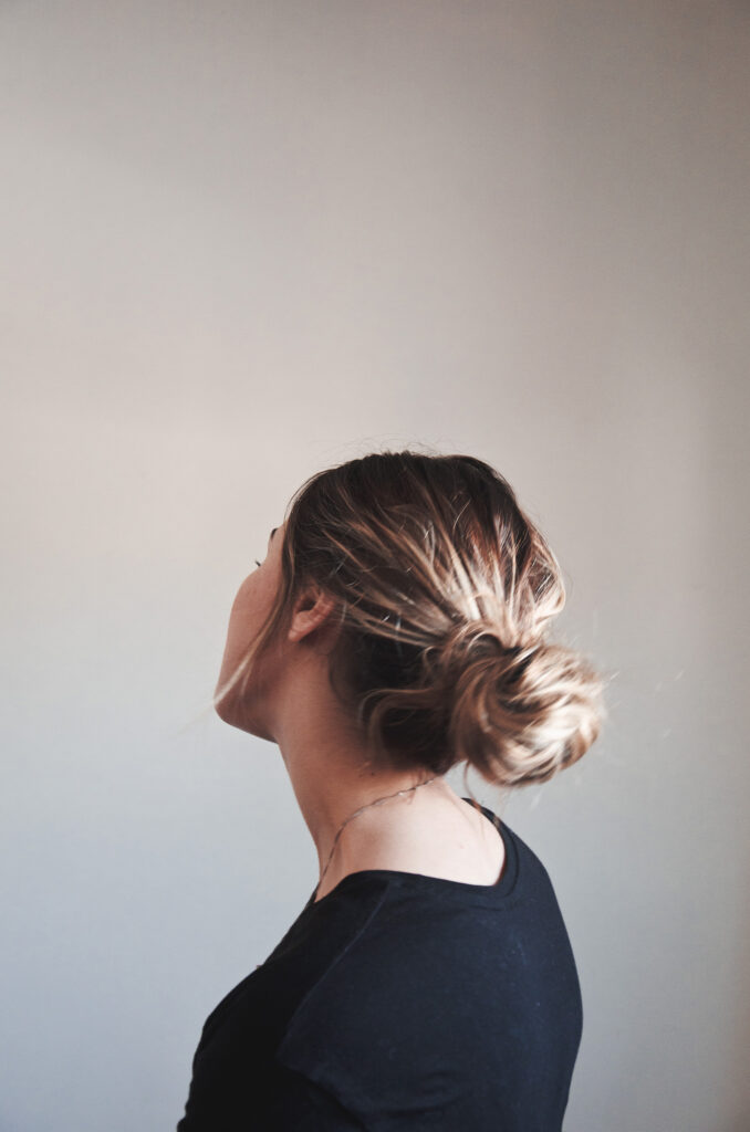 Darian Rojas's profile view showcases a neatly tied bun a delicate necklace and a dark-colored top against a muted background.